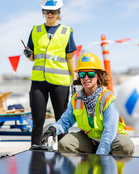 Two women in safety vests and hard hats inspect solar panels at a construction site
