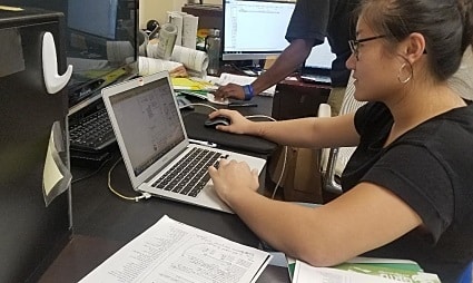 Woman wearing glasses working at laptop in office with papers and multiple monitors visible