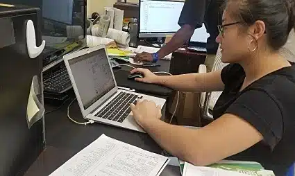 Woman wearing glasses working at laptop in office with papers and multiple monitors visible