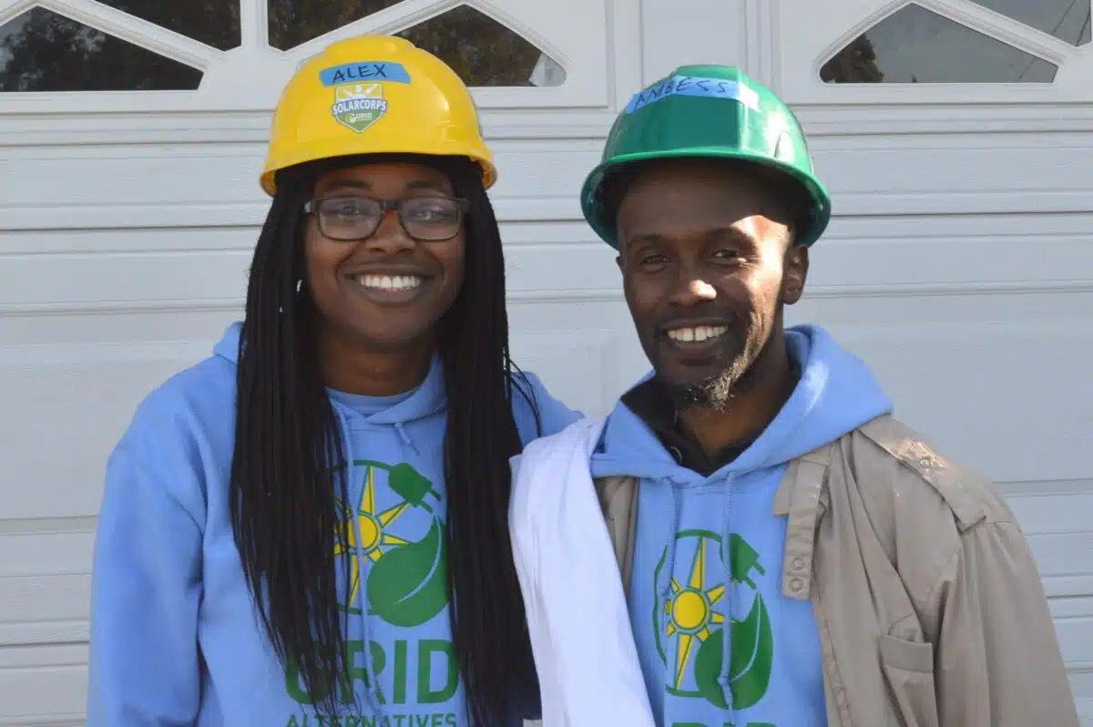 Two people wearing blue hoodies and construction hard hats smile in front of a white garage door