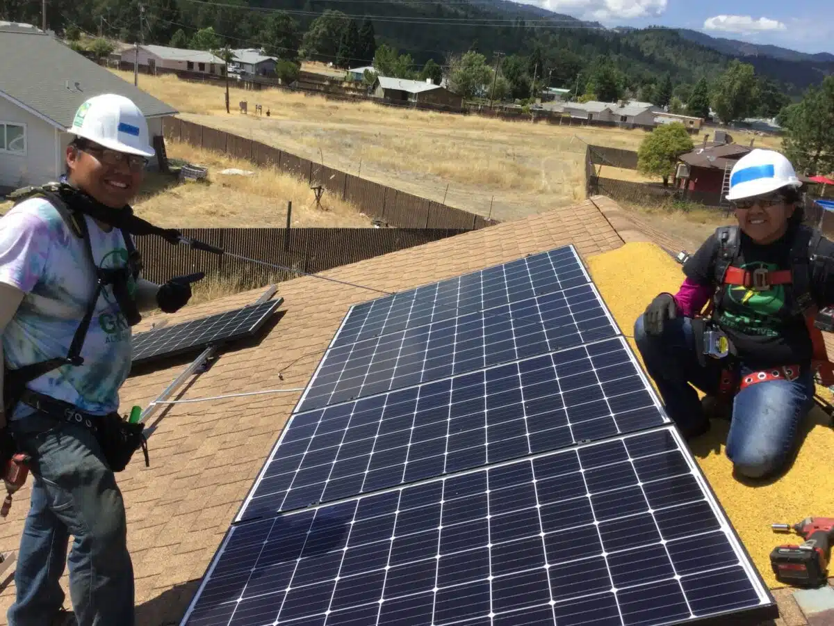 Two workers in safety gear installing solar panels on a residential roof in a mountain valley