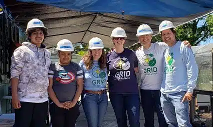 Six people wearing white hard hats and GRID-branded shirts stand together under a tent at an outdoor event