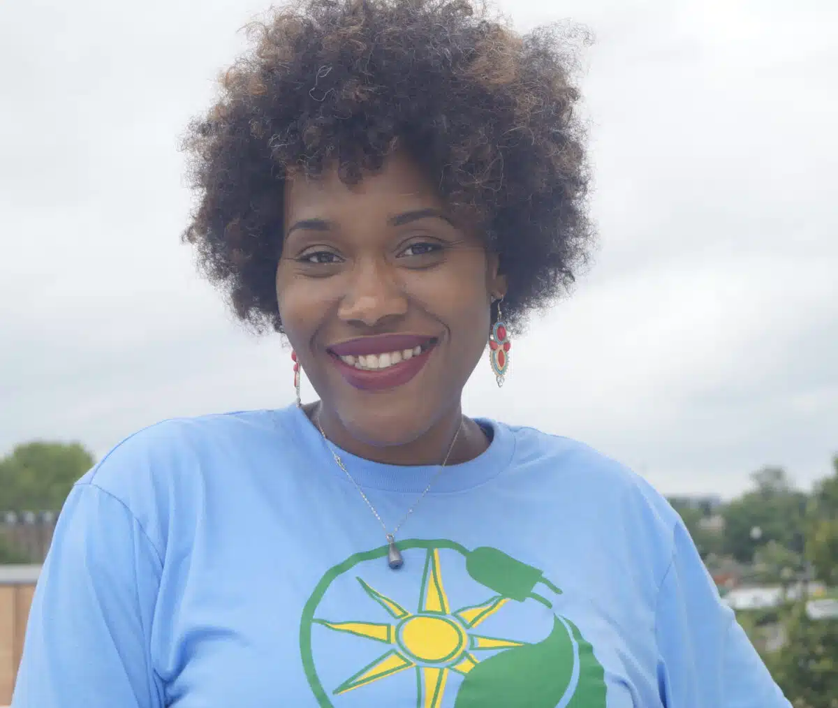 Woman with curly hair smiling outdoors, wearing light blue shirt with green and yellow sun logo