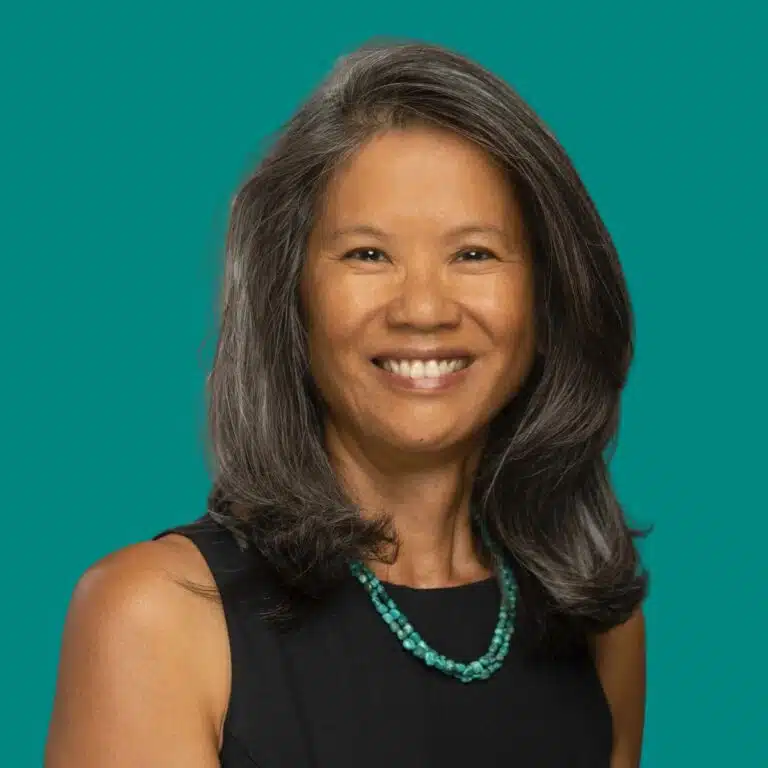 Woman with long dark hair wearing a black sleeveless top and turquoise beaded necklace, smiling against a teal background