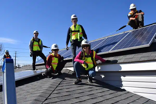 Four young people stand or crouch on top of a roof while wearing hard hats and harnesses