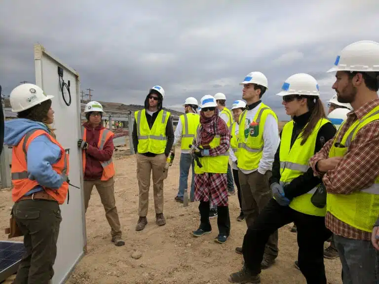 A group of people wearing yellow safety vests listens to a GRID construction staff person as she shows them the back of a solar panel