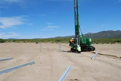 Green drilling rig on sandy desert ground with blue PVC pipes and mountains in background