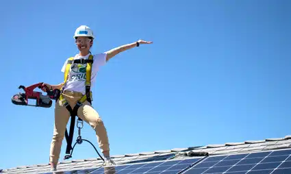 Construction worker in safety gear standing on solar panels, pointing outward against blue sky