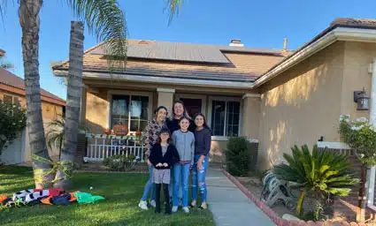 Family of five standing on walkway in front of single-story house with solar panels