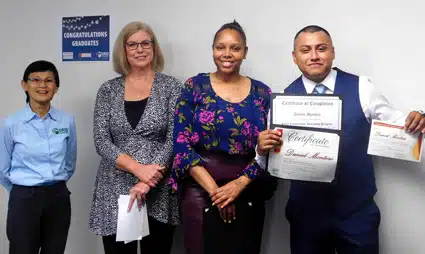 Four people posing together with certificates at a graduation event, congratulations sign visible on wall behind them