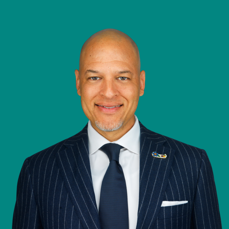 Professional headshot of a bald man in a dark pinstriped suit with navy tie against turquoise background