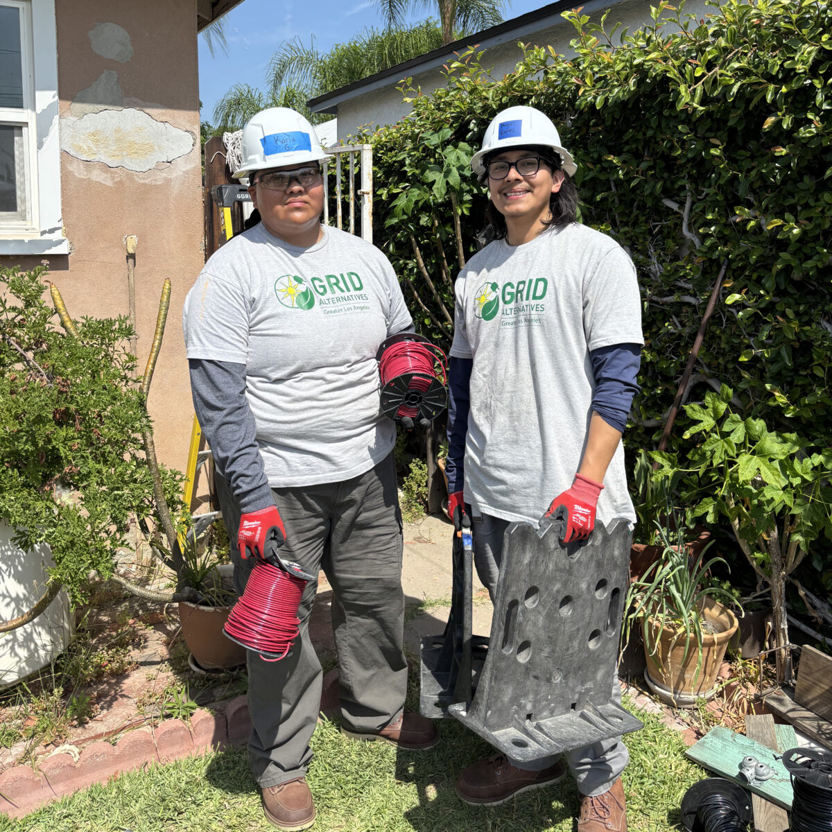 Karla and a fellow IBT 200 trainee in GRID Alternatives shirts and hard hats hold spools of red wire and solar mounting equipment outside a home.