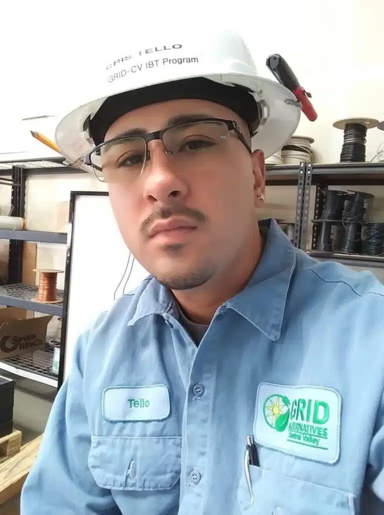 Worker named Tello wearing GRID Alternatives hard hat, safety glasses, and blue work shirt in warehouse setting