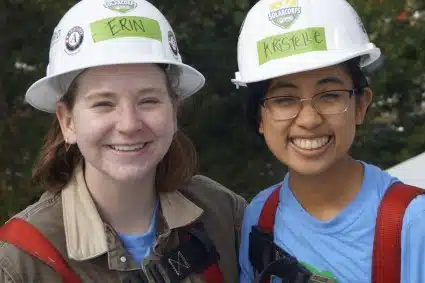 Two smiling young women in hard hats labeled "CKMI" and "KSCLF" wearing safety vests outdoors