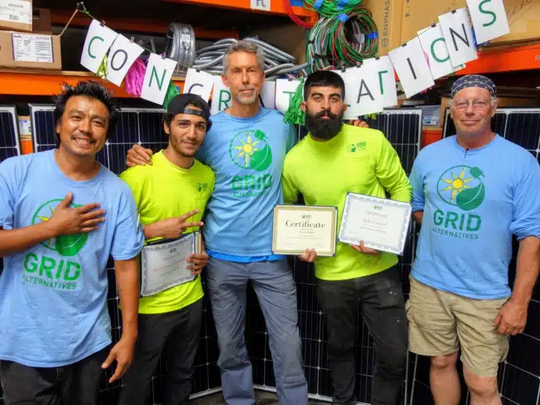 Five GRID Alternatives team members holding certificates in front of solar panels and "Congratulations" banner