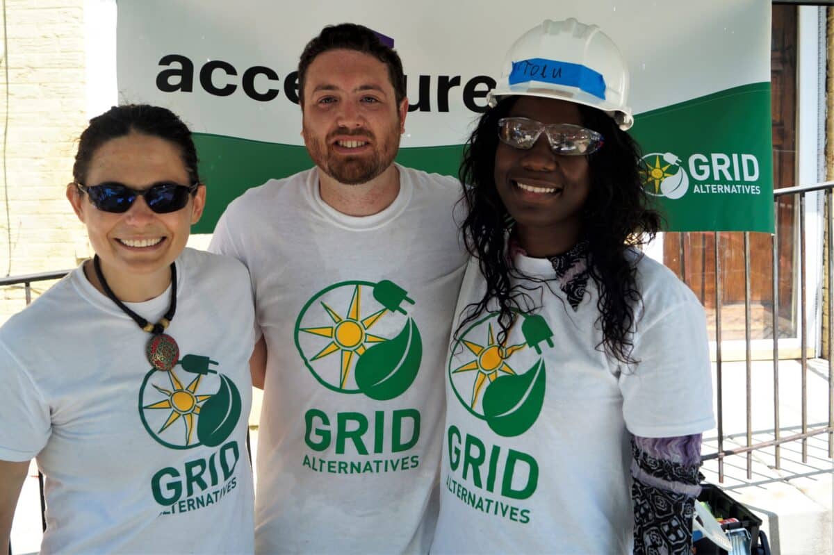 Three people wearing Grid Alternatives t-shirts stand together at an outdoor event, smiling at the camera