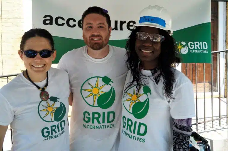 Three people wearing Grid Alternatives t-shirts stand together at an outdoor event, smiling at the camera