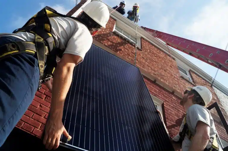 Workers installing solar panels on a brick rooftop with safety harnesses and hard hats