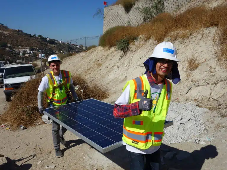 Two construction workers in hard hats and safety vests holding a blue solar panel on a hillside