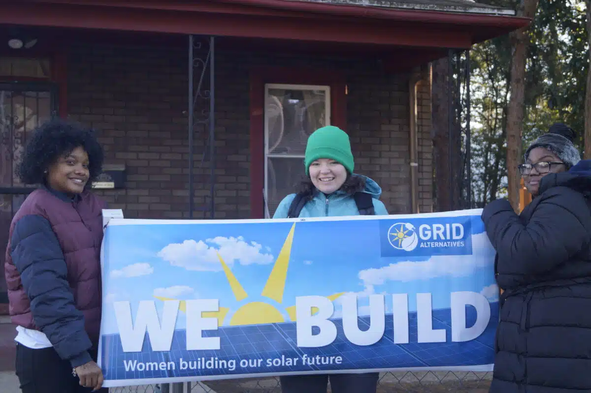 Three women holding a "We Build" banner for GRID Alternatives' solar energy program outside a brick building