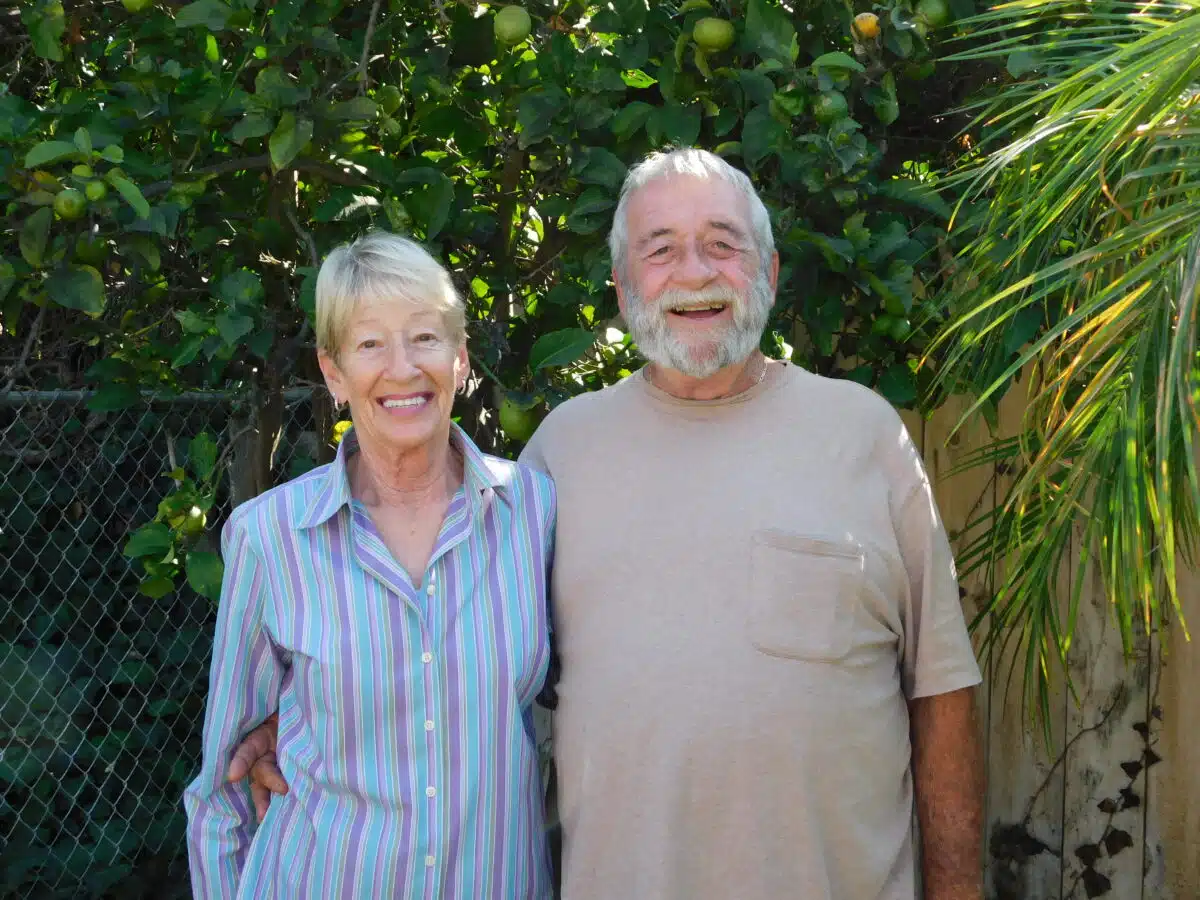 Two smiling older adults standing together in a garden with lush green foliage and palm fronds behind them