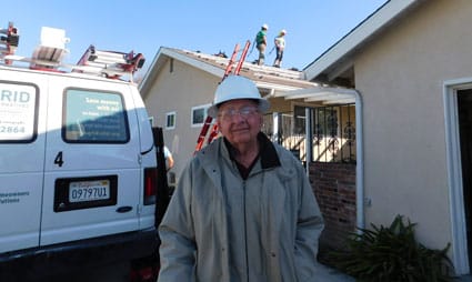 Contractor in hard hat standing in front of house with work van and roofing crew visible