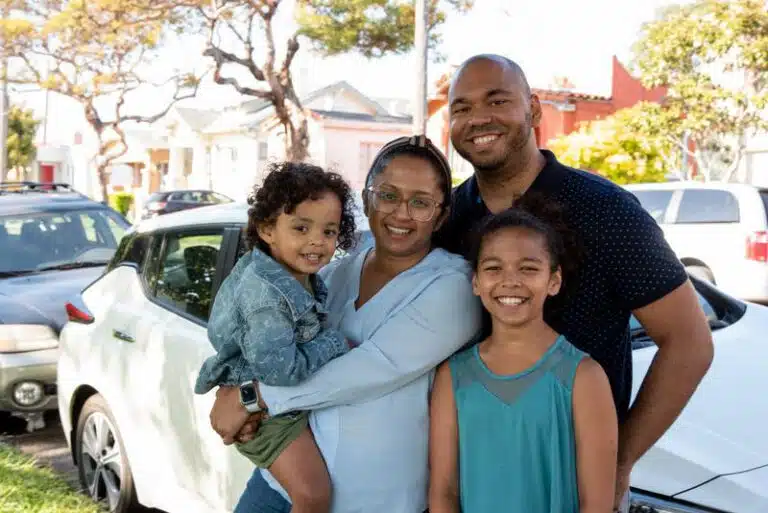 Happy family of four standing in front of white electric vehicle in sunny residential neighborhood