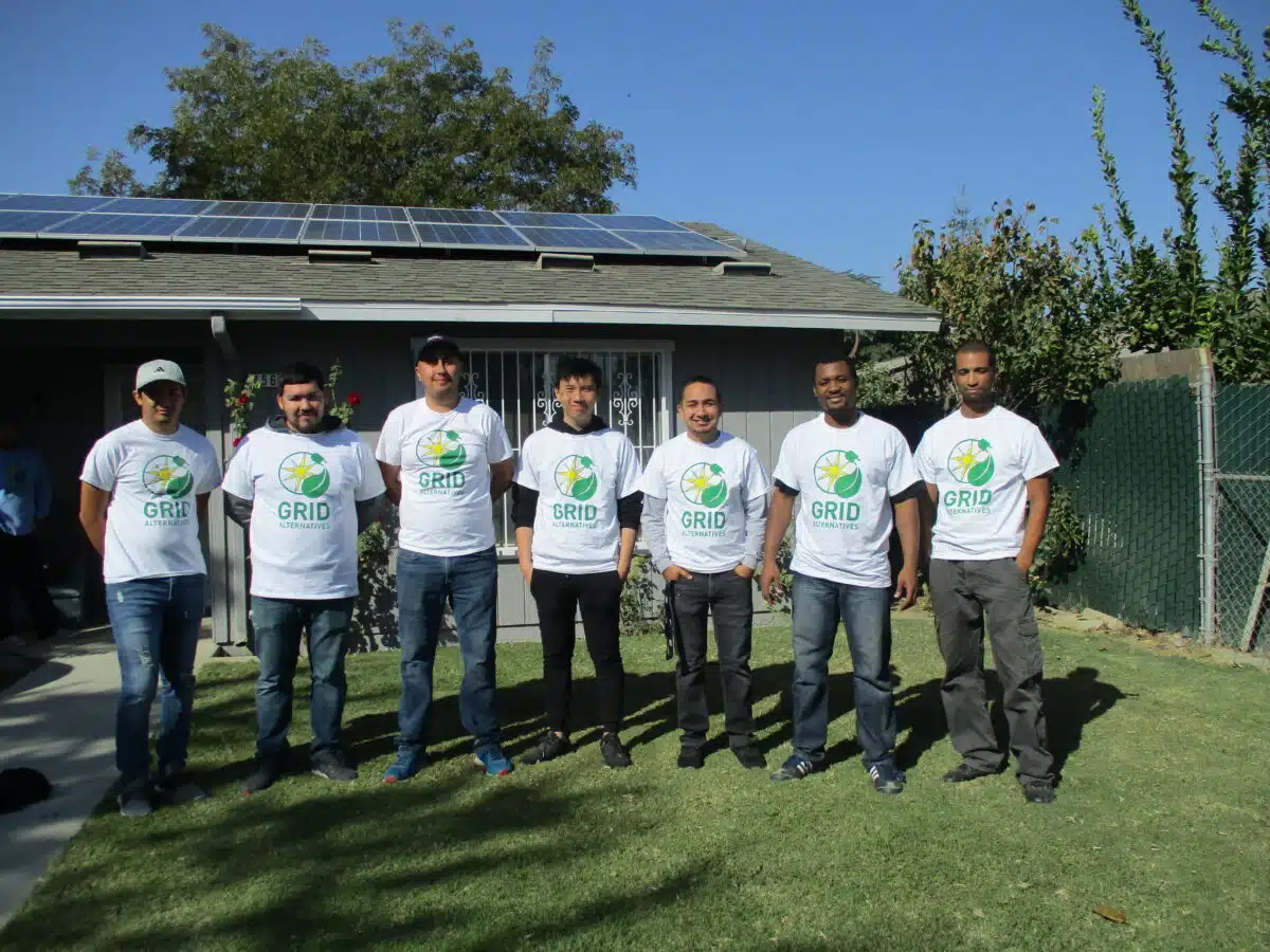 Seven people wearing GRID Alternatives t-shirts stand in front of a house with solar panels on the roof