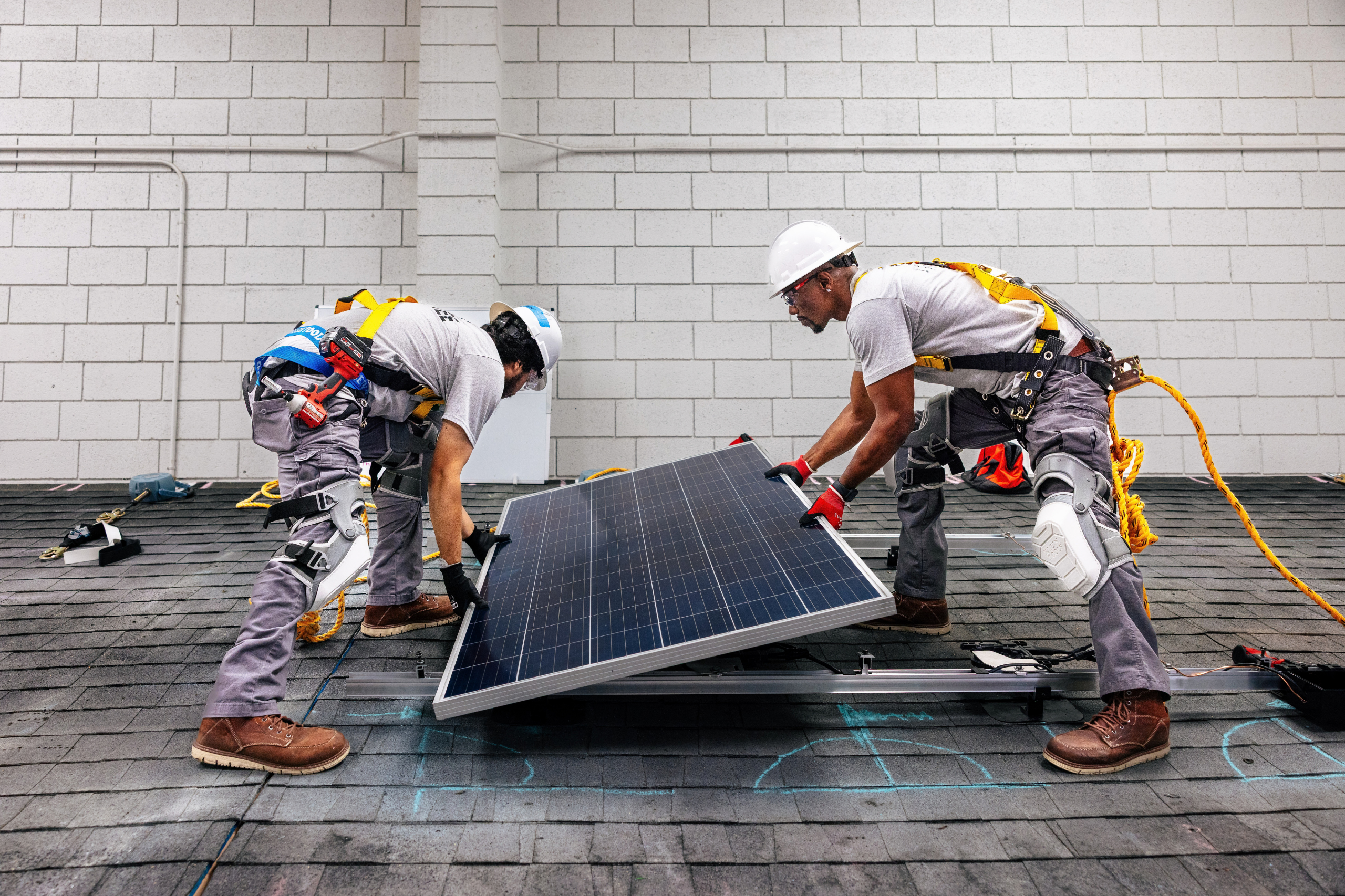Two workers install a solar panel on a residential rooftop, wearing hard hats and safety harnesses