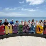 Group of eleven people posing with colorful "Ensenada" letters at a coastal overlook with ocean and rocks in background