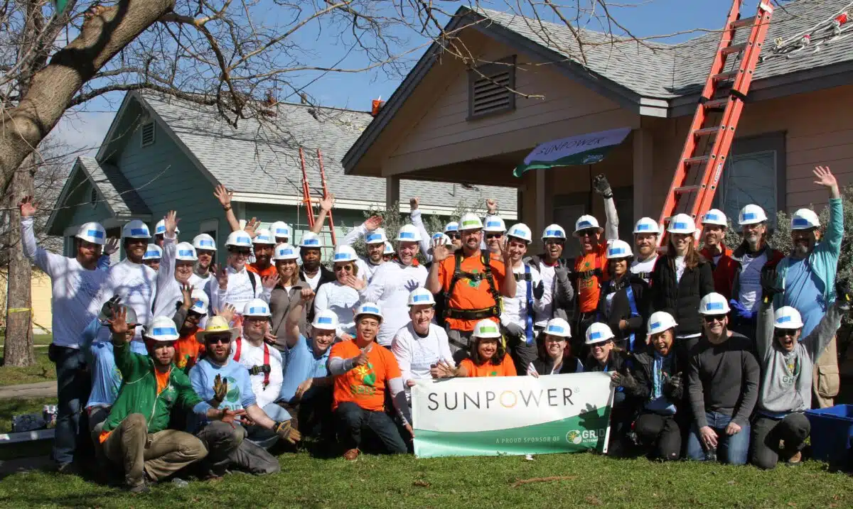 Large group of volunteers in hard hats holding SunPower banner in front of houses under renovation