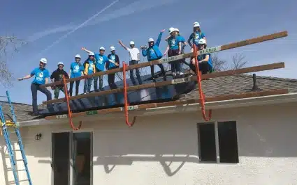 Large group of volunteers in blue shirts installing roof framing on a white building