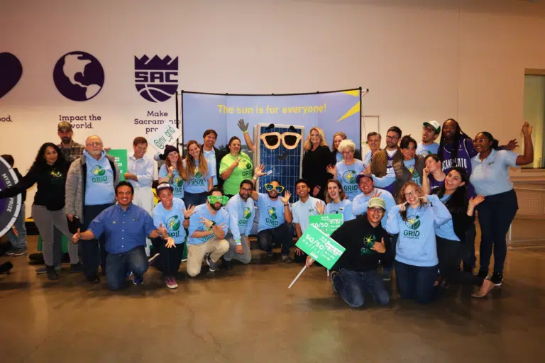 Large group of people wearing green and blue GRID shirts posing together indoors with solar panel banner, "The sun is for everyone!