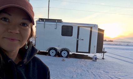 Woman in cap smiling at camera with white travel trailer on snowy beach at sunset
