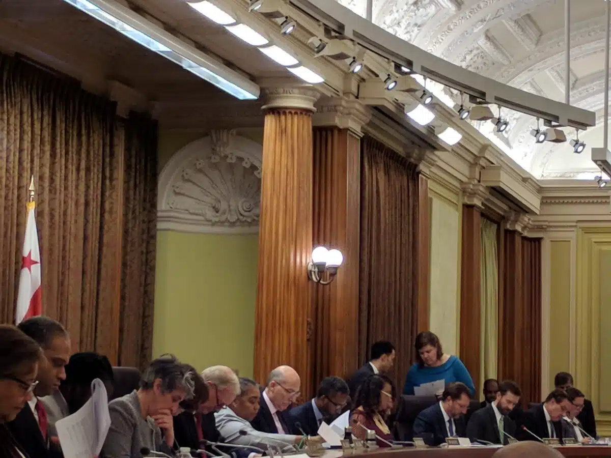 Government officials seated at a formal meeting table in an ornate chamber with columns and a D.C. flag visible