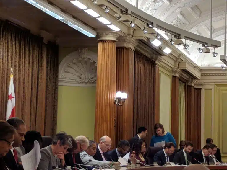 Government officials seated at a formal meeting table in an ornate chamber with columns and a D.C. flag visible