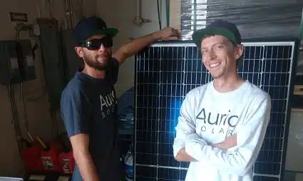 Two men in Auric Solar shirts stand beside solar panels in an indoor setting