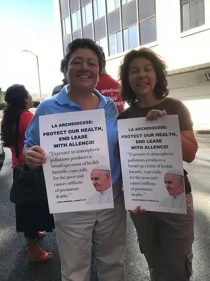 Two individuals supporting action by the Archdiocese hold up signs that say, "LA Archdiocese: Protect our health, end the lease with AllenCo"
