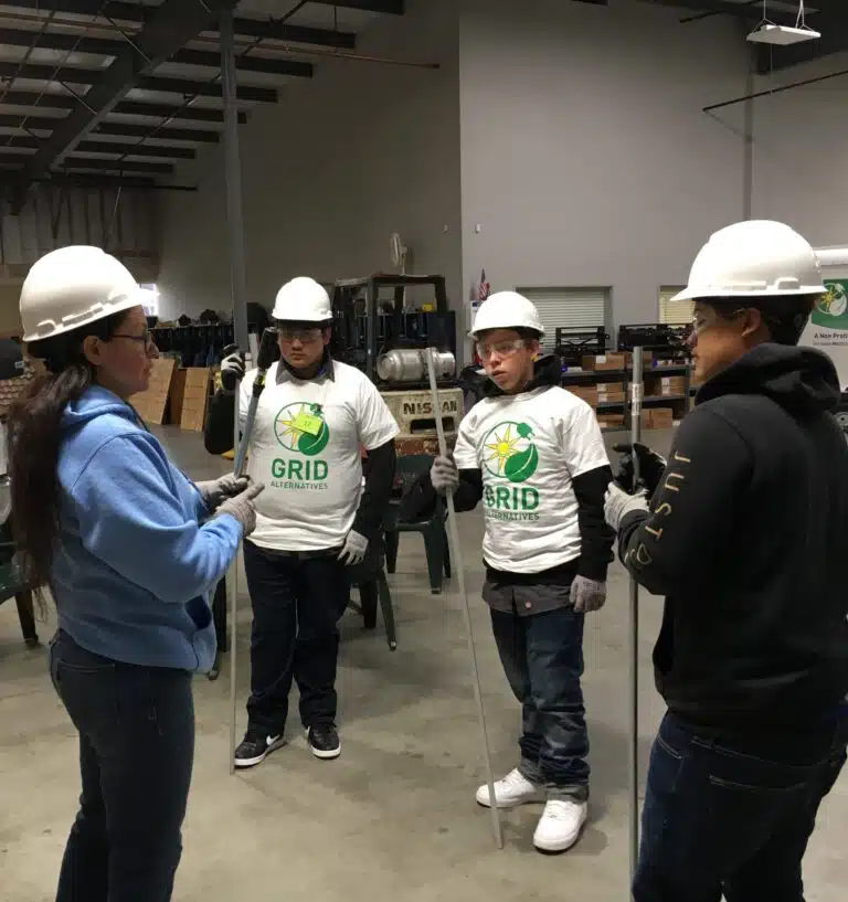 Five people wearing hard hats and work gloves stand in a warehouse, three wearing GRID Alternatives branded shirts during what appears to be a training or work session