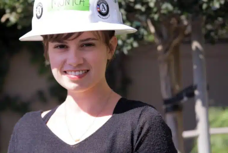 Smiling young person wearing white hard hat with logos and dark shirt