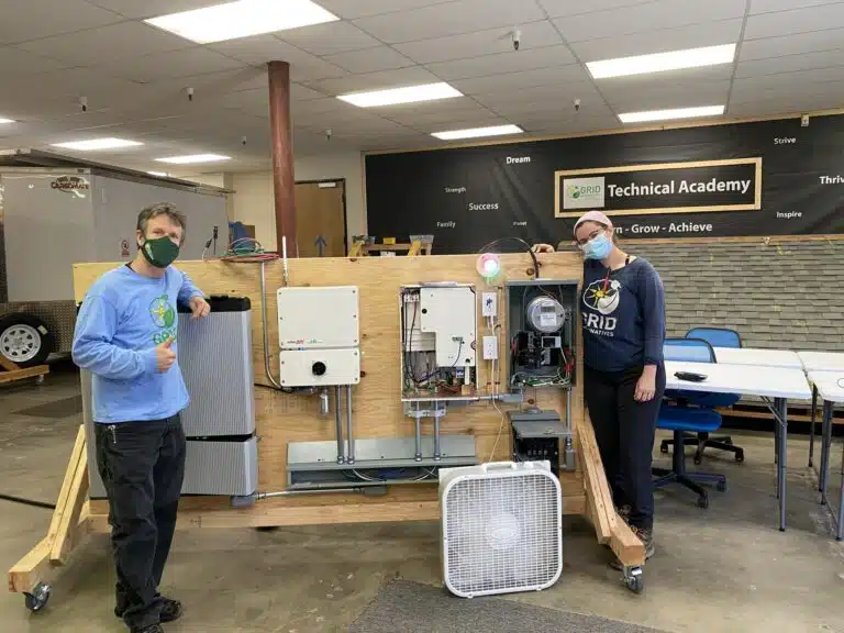 Two people displaying a wooden-framed HVAC system demonstration unit at GRID Technical Academy