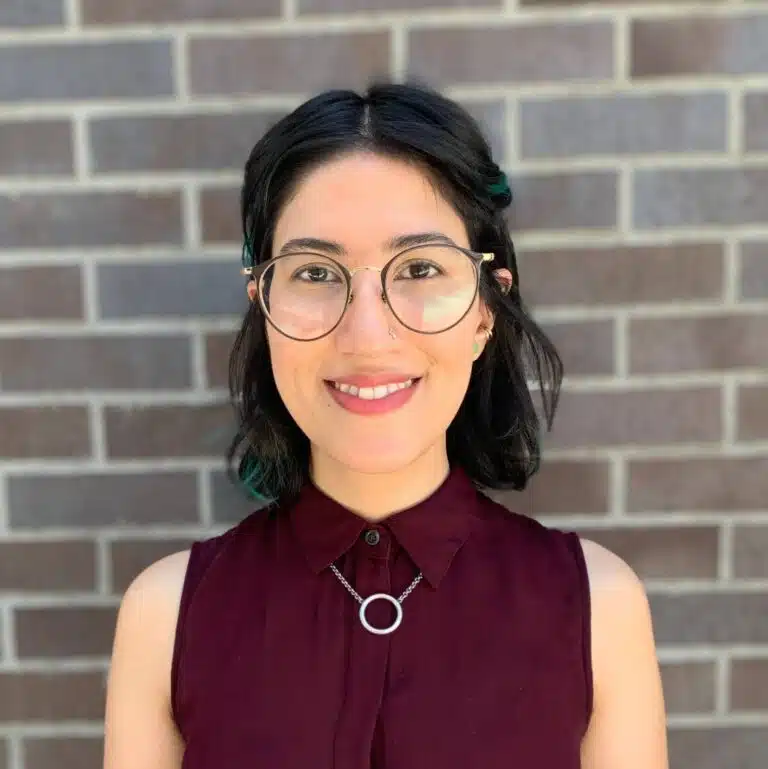 Woman with dark hair and glasses wearing a maroon sleeveless shirt against a brick wall