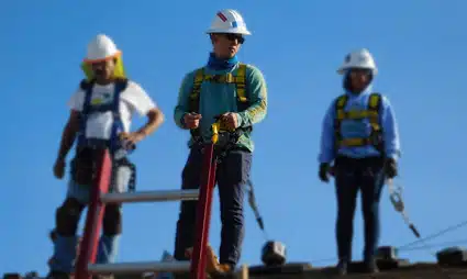 Three construction workers in safety gear and hard hats stand against a blue sky