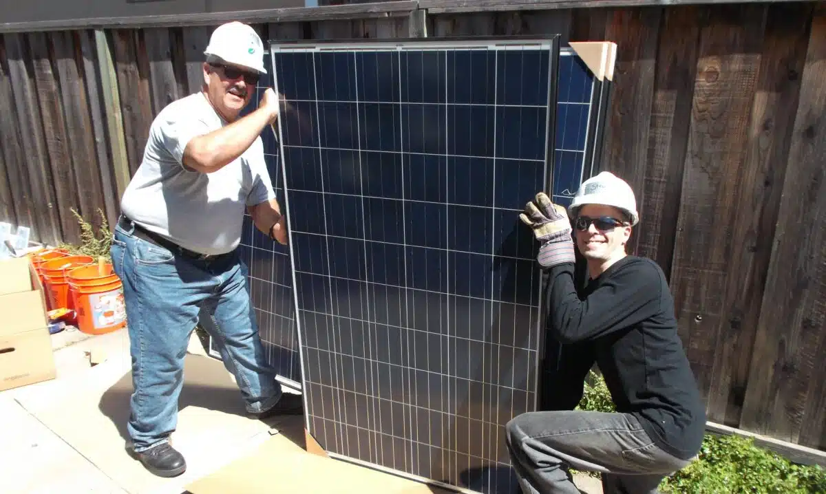 Two workers in hard hats and safety gear installing a large solar panel against a wooden fence