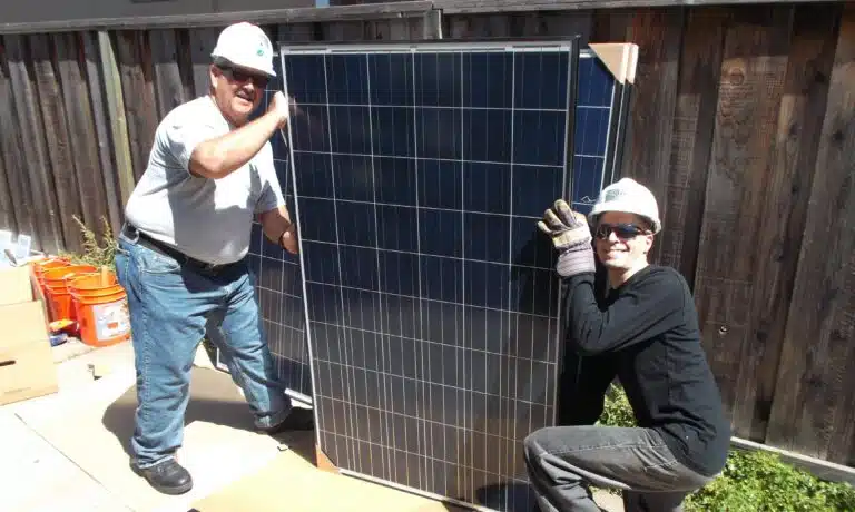 Two workers in hard hats and safety gear installing a large solar panel against a wooden fence