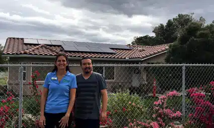 Two people standing in front of a house with solar panels and flowering plants along a fence