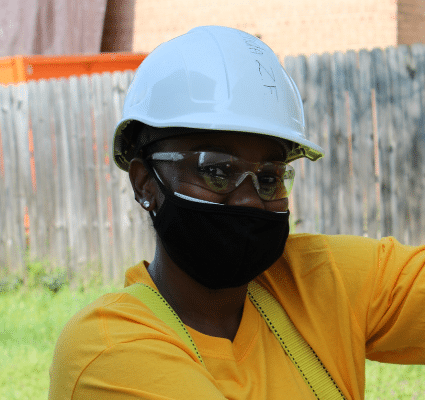 Construction worker wearing blue hard hat, black mask, yellow shirt, and safety glasses