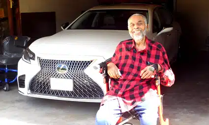 Older man in red plaid shirt posing with cane in front of white Lexus car