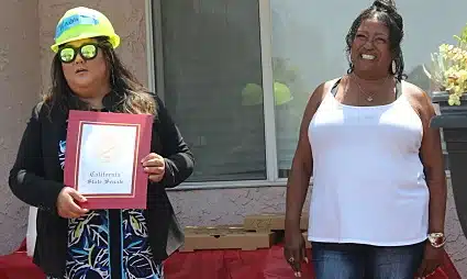 Smiling, a white-shirted homeowner accepts a certificate from another person wearing a safety helmet and dress