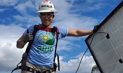 Worker named Natalie in hard hat and safety harness installing solar panel against blue sky